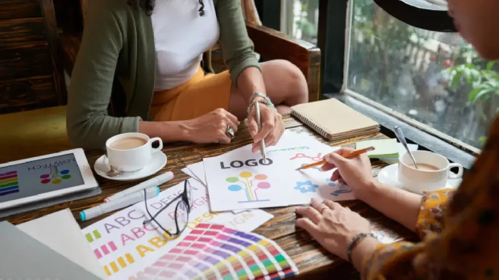 Two individuals collaborating on logo design and branding concepts, surrounded by colorful tools, sketches, coffee cups, and a creative workspace.