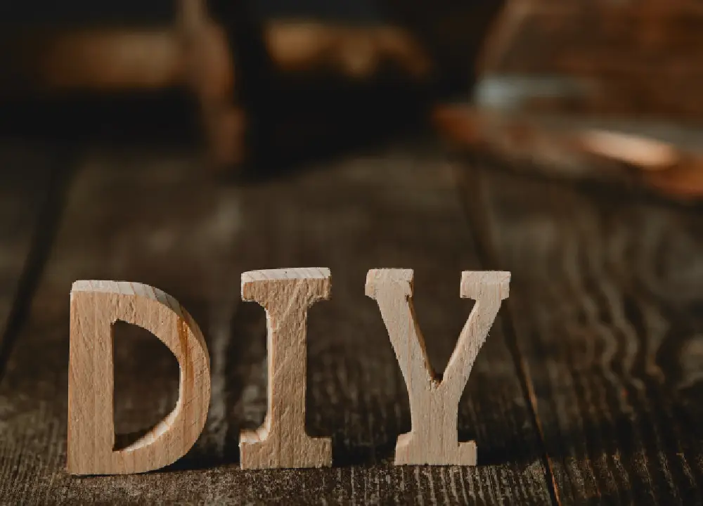 Close-up of a DIY sign on a dark wooden table, with paintbrushes, paint, and logs in the background, highlighting the handmade and rustic feel.

